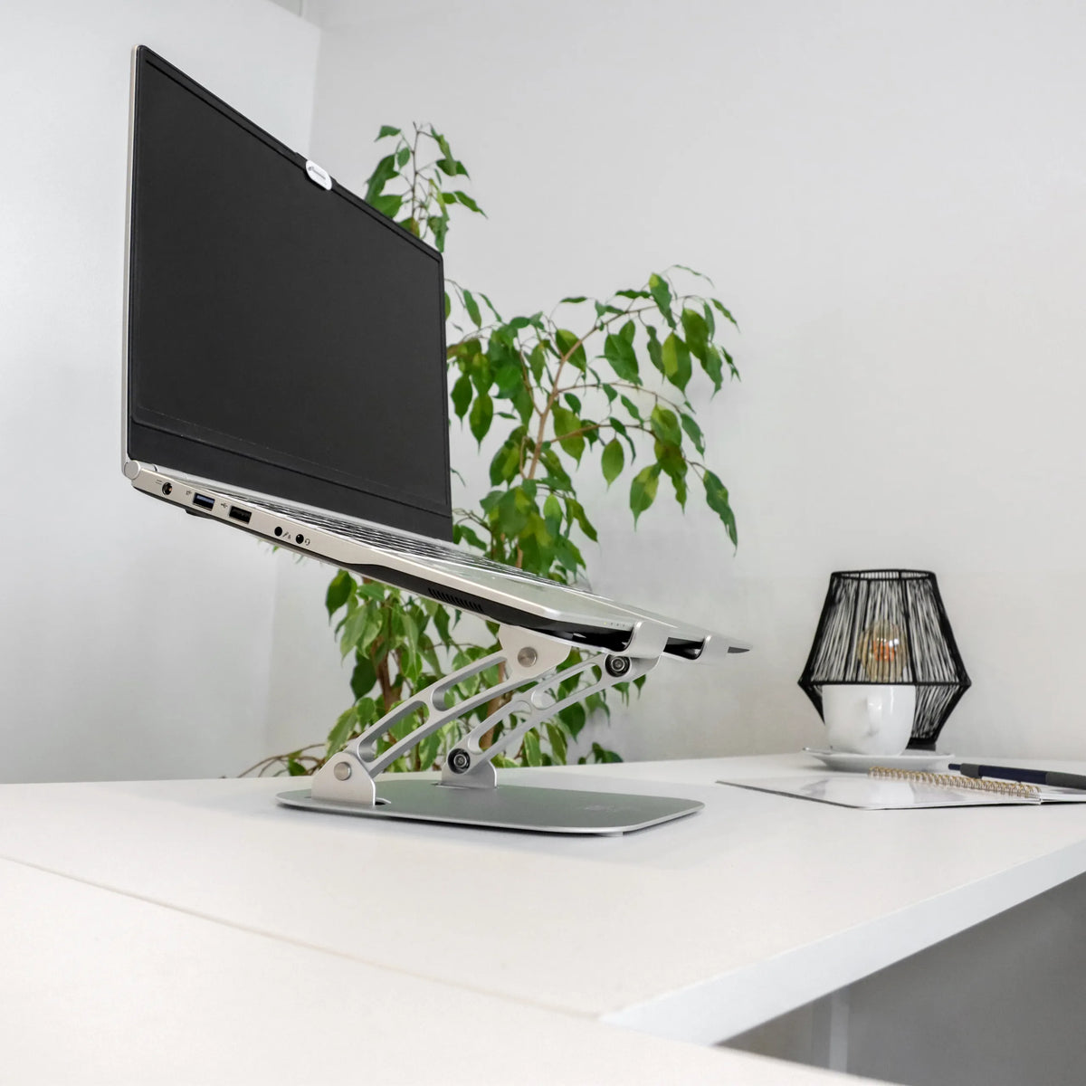 Soporte para portátil LAPTOP STAND plateado con estructura regulable en altura y portátil sobre un escritorio blanco de hjh OFFICE