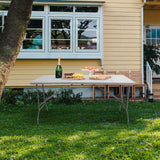Mesa plegable blanca EASY UP MULTI II de 152 x 70 cm en un césped frente a una casa de hjh OFFICE