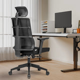 Silla de oficina profesional SPINIO PRO negra con reposacabezas y reposabrazos delante de un escritorio de hjh OFFICE